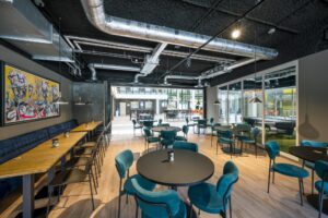 Moderne, industrieel vormgegeven kantine aan de Thomas R. Malthusstraat met turquoise stoelen, houten vloeren en zicht op een lichte binnenruimte.